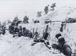 Photograph, 4th Division troops shelter behind a concrete wall while others advance off the beach.